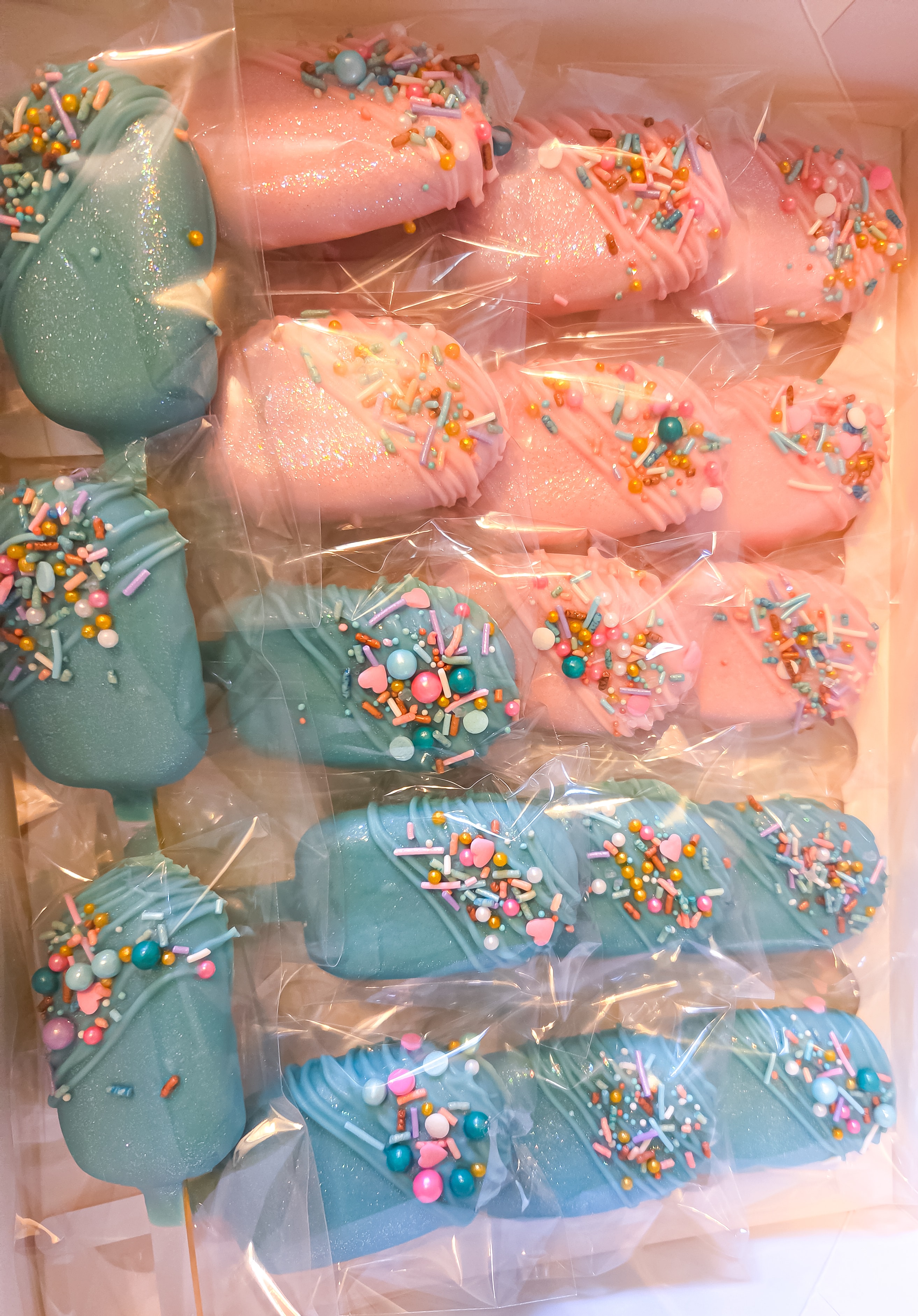 Pink and teal chocolate-covered cakesicles with sprinkles, packaged in cellophane